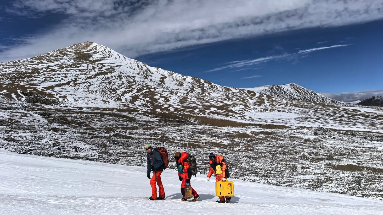 Three people walking up a snow covered hill, surrounded by mountains, with blue skies and white clouds..