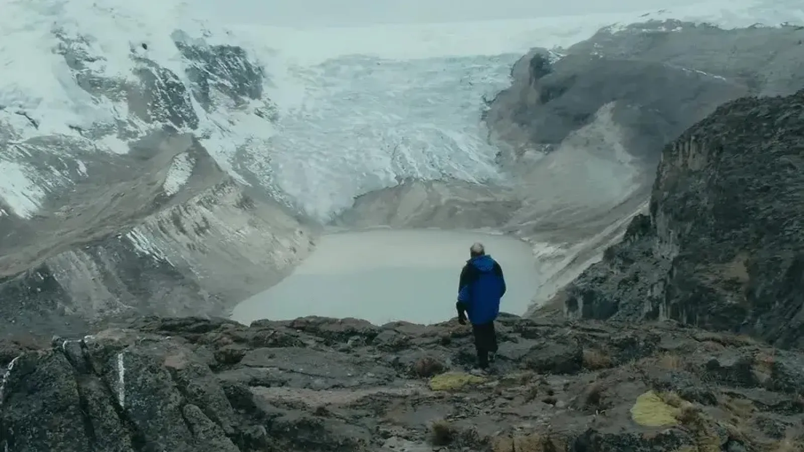 Person in blue jacket standing on rocky ground, observing a glacier and a glacial lake in a vast, mountainous landscape.