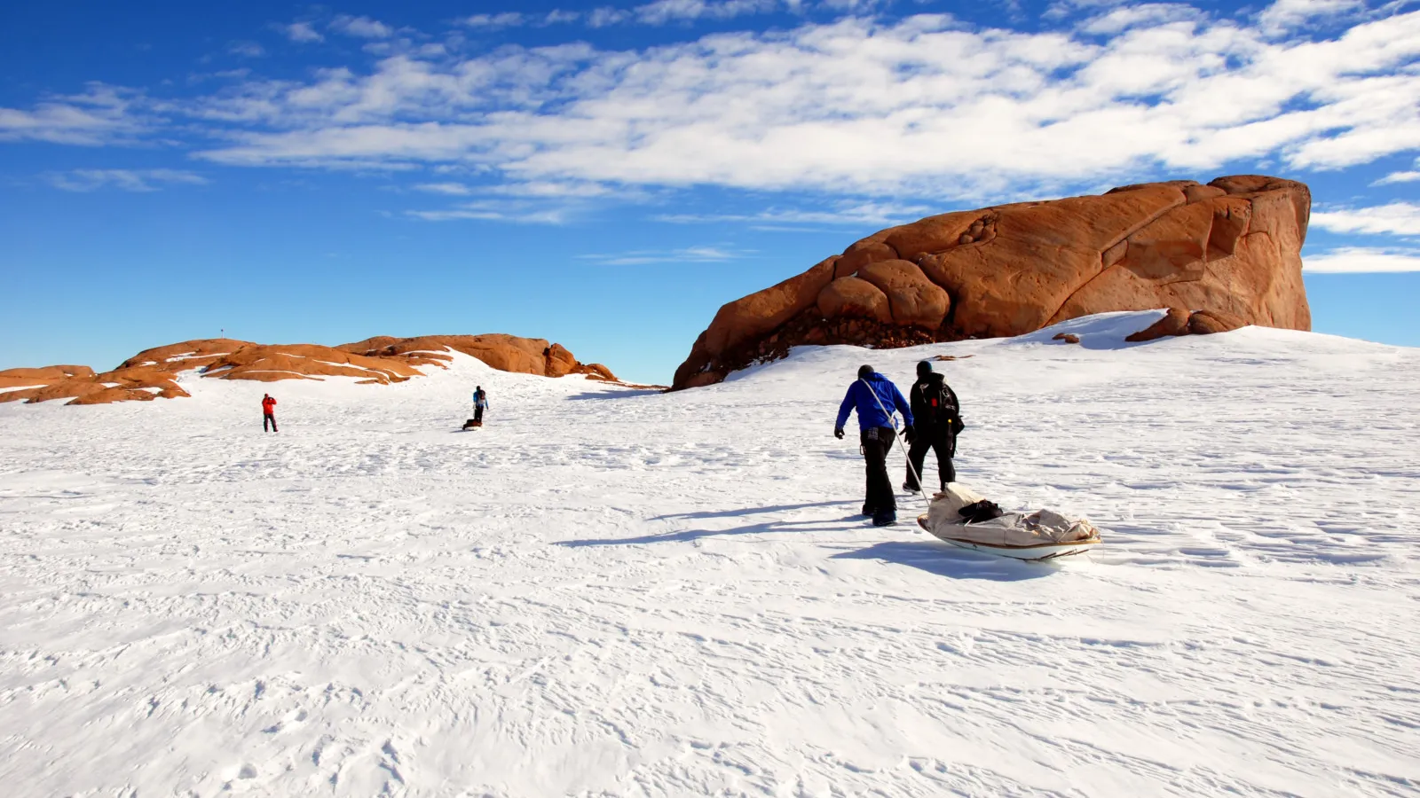 People participate in snow activities near a large sandstone rock formation under a clear blue sky.
