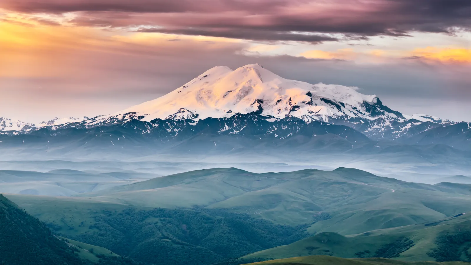 view of Elbrus mount in the morning at sunrise with a dramatic cloudy sky and foggy green highland meadows. Summer mountain landscape. North Caucasus, Karachay-Cherkess Republic, Russia