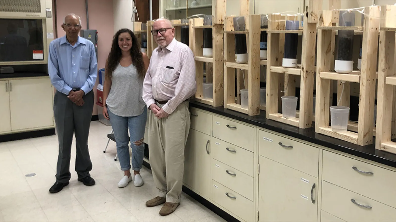 Three individuals standing in a laboratory with shelves containing scientific samples behind them.