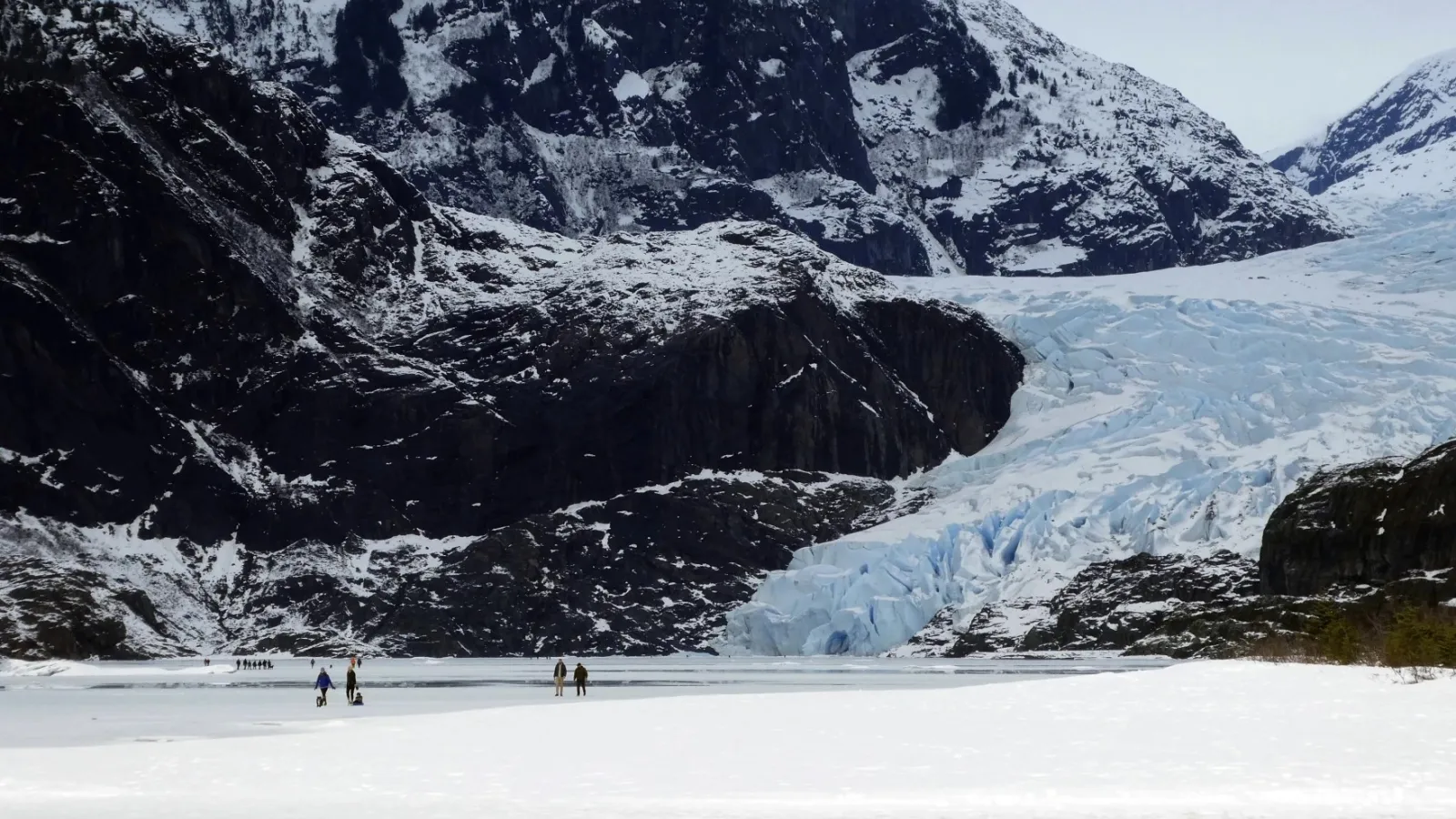 People walking near a Glacier, with snow-covered mountains in the background.