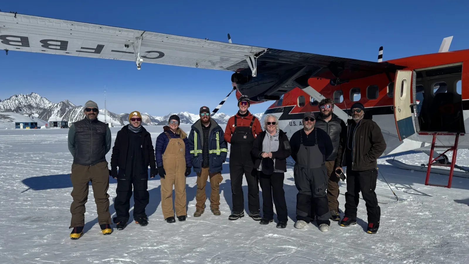 Group of ten people standing in front of a red and white plane on a snowy landscape with mountains in the background.