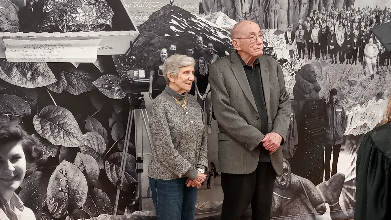 Two individuals standing in front of a large black and white photo collage featuring historical images and photographs of people.