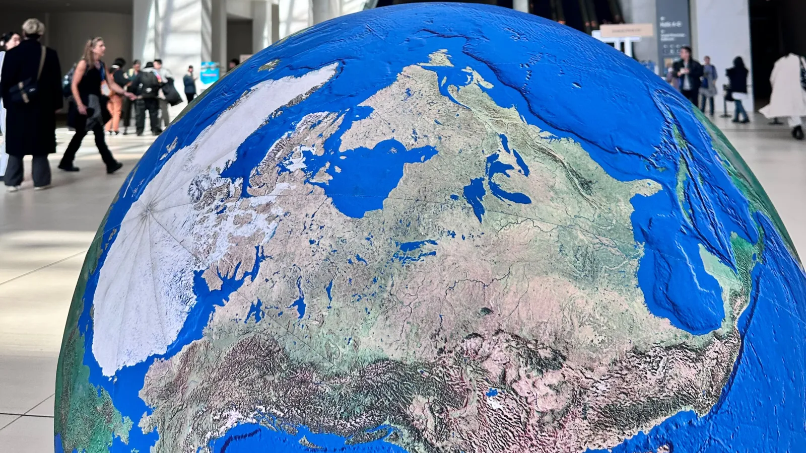 A large, detailed globe model  positioned in the foreground of a spacious convention center lobby. People walk and gather in the background near escalators.