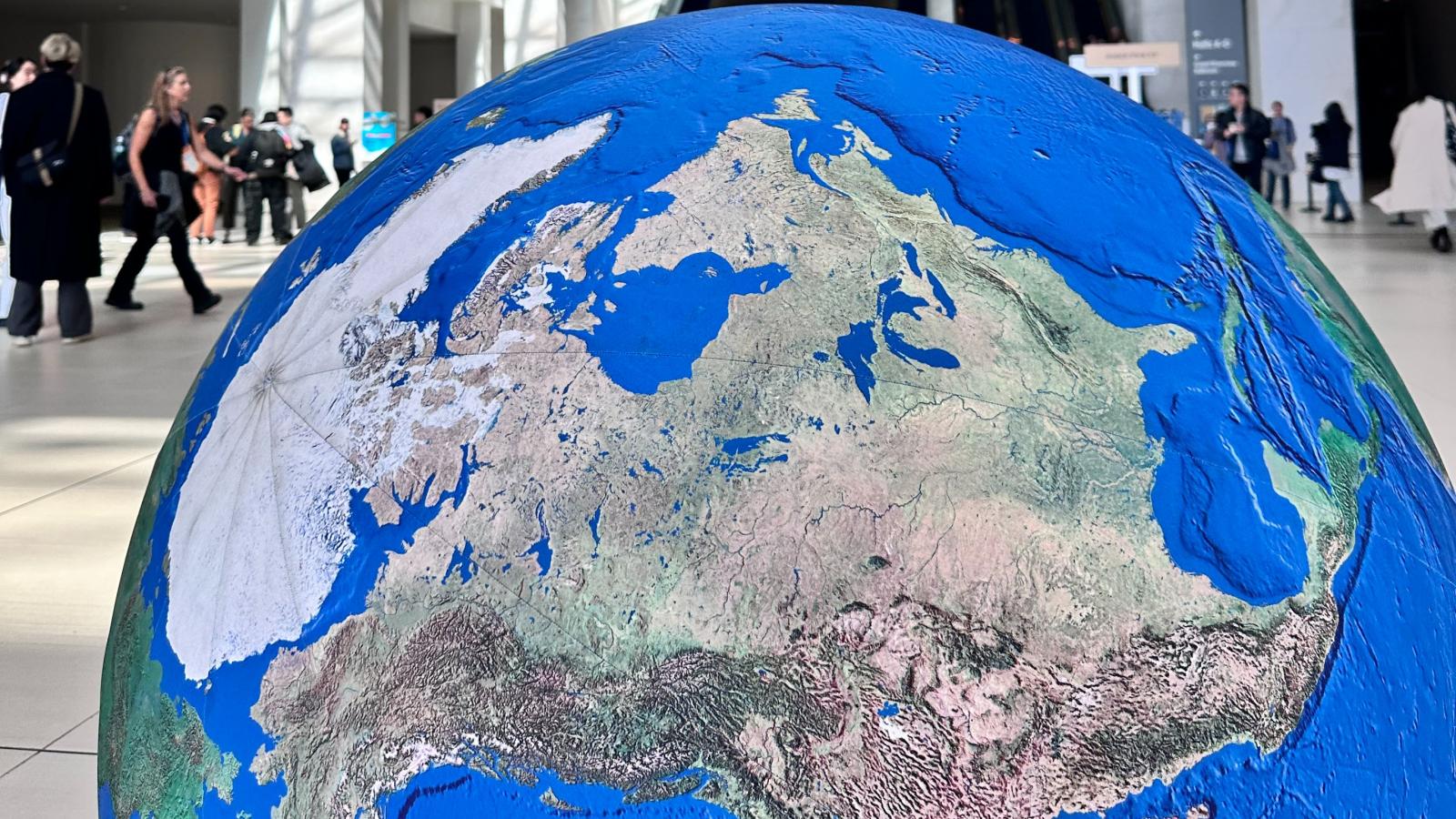 A large, detailed globe model  positioned in the foreground of a spacious convention center lobby. People walk and gather in the background near escalators.