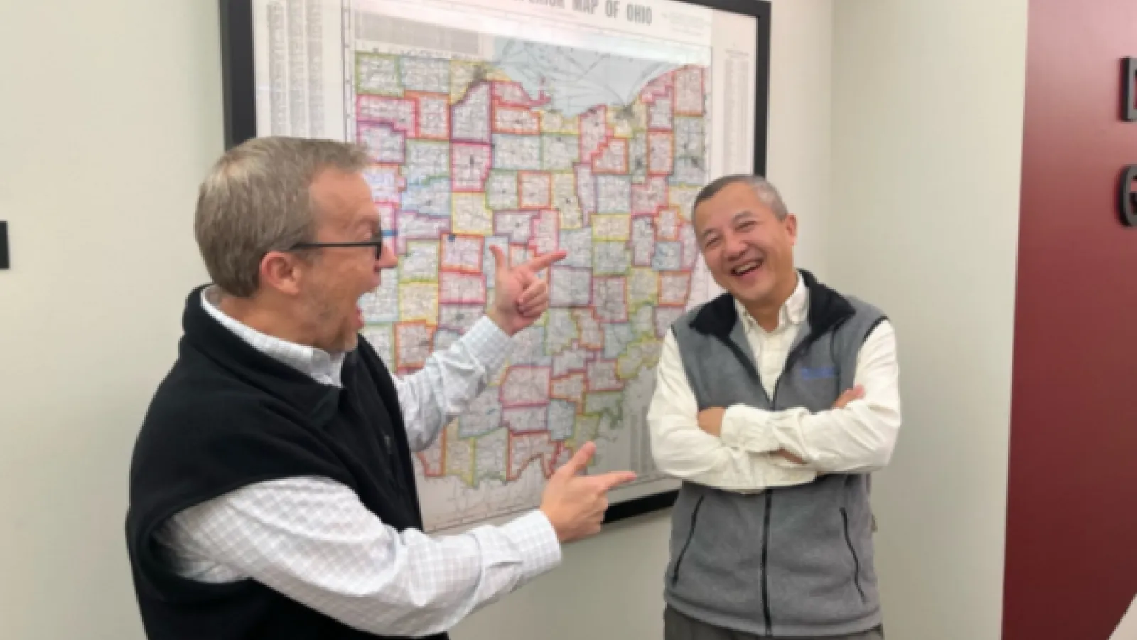 Two individuals laugh and chat in front of a framed map of Ohio; one points playfully toward the other.