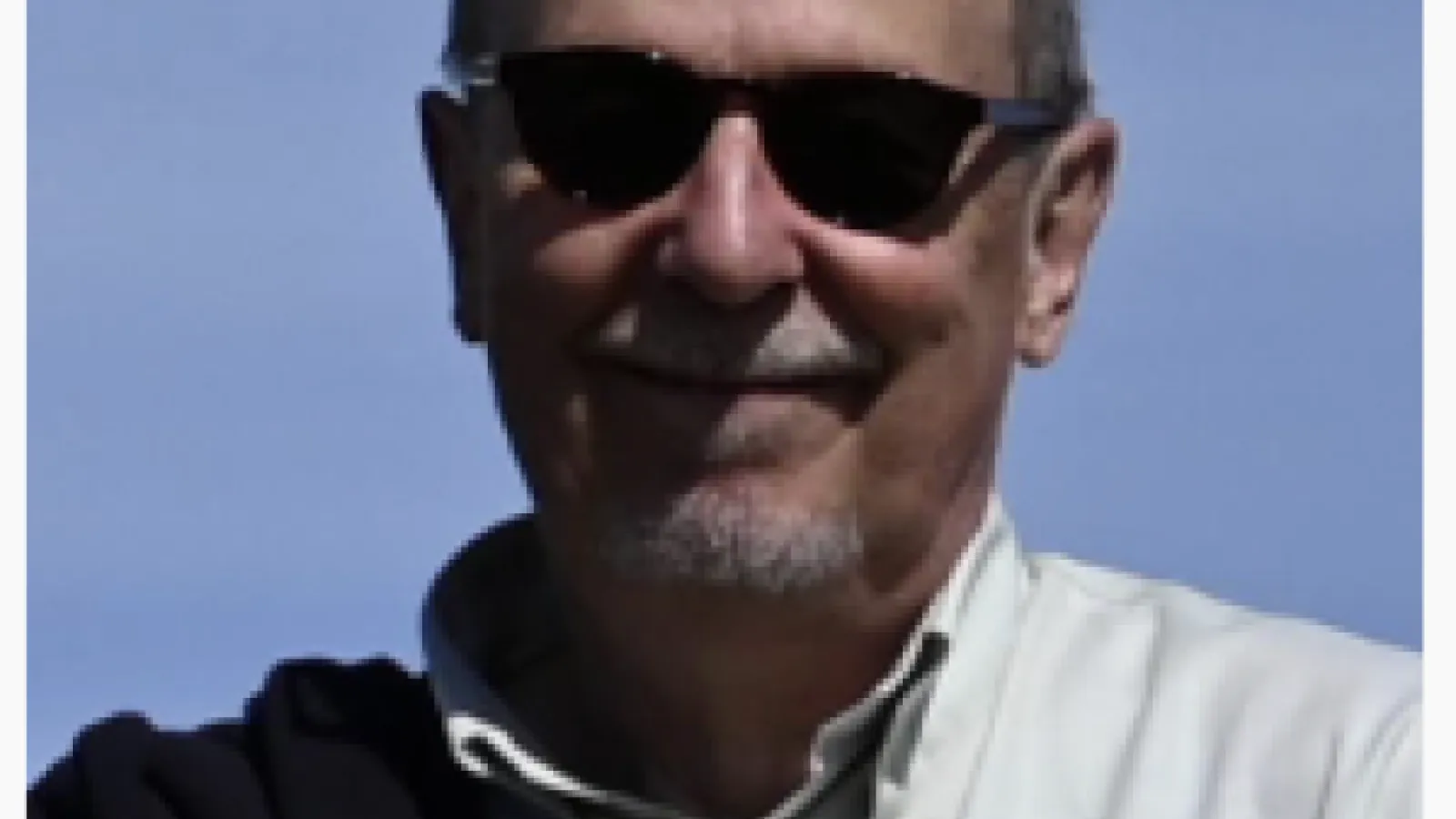 A man with a beard wearing dark sunglasses and a light-colored button-down shirt. He is smiling.