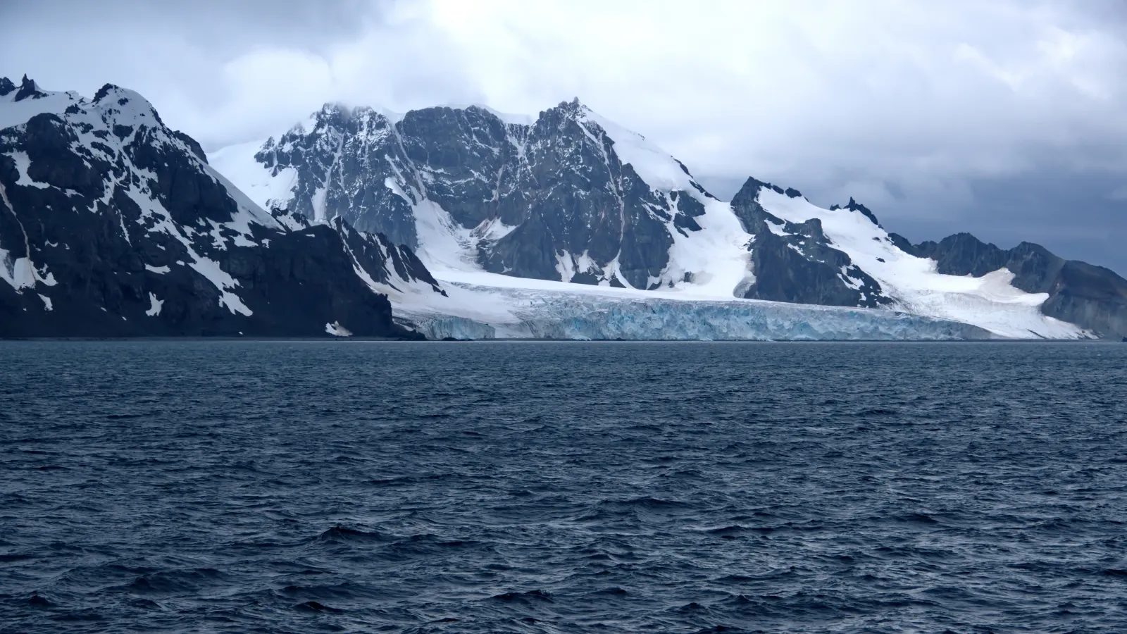 Snow-covered mountains and glacier going into the sea under a cloudy sky.