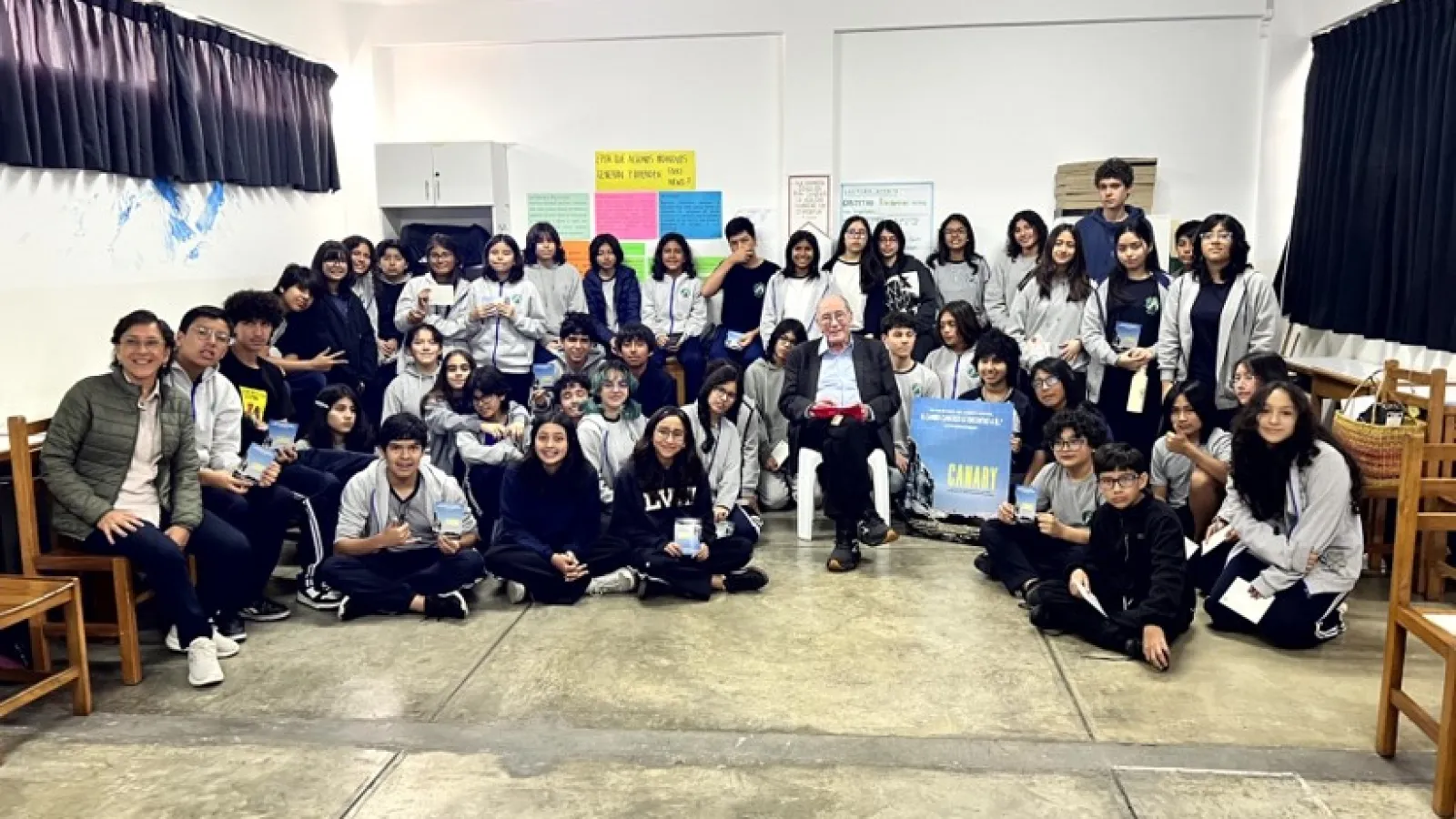 Group photo of high school students and teachers gathered around a seated guest speaker holding a CANARY movie poster.
