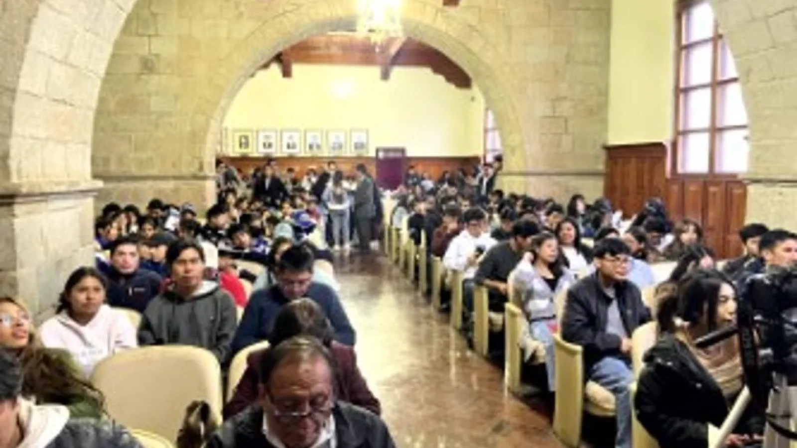 people fill rows of seats in a stone-arched lecture hall before a presentation.