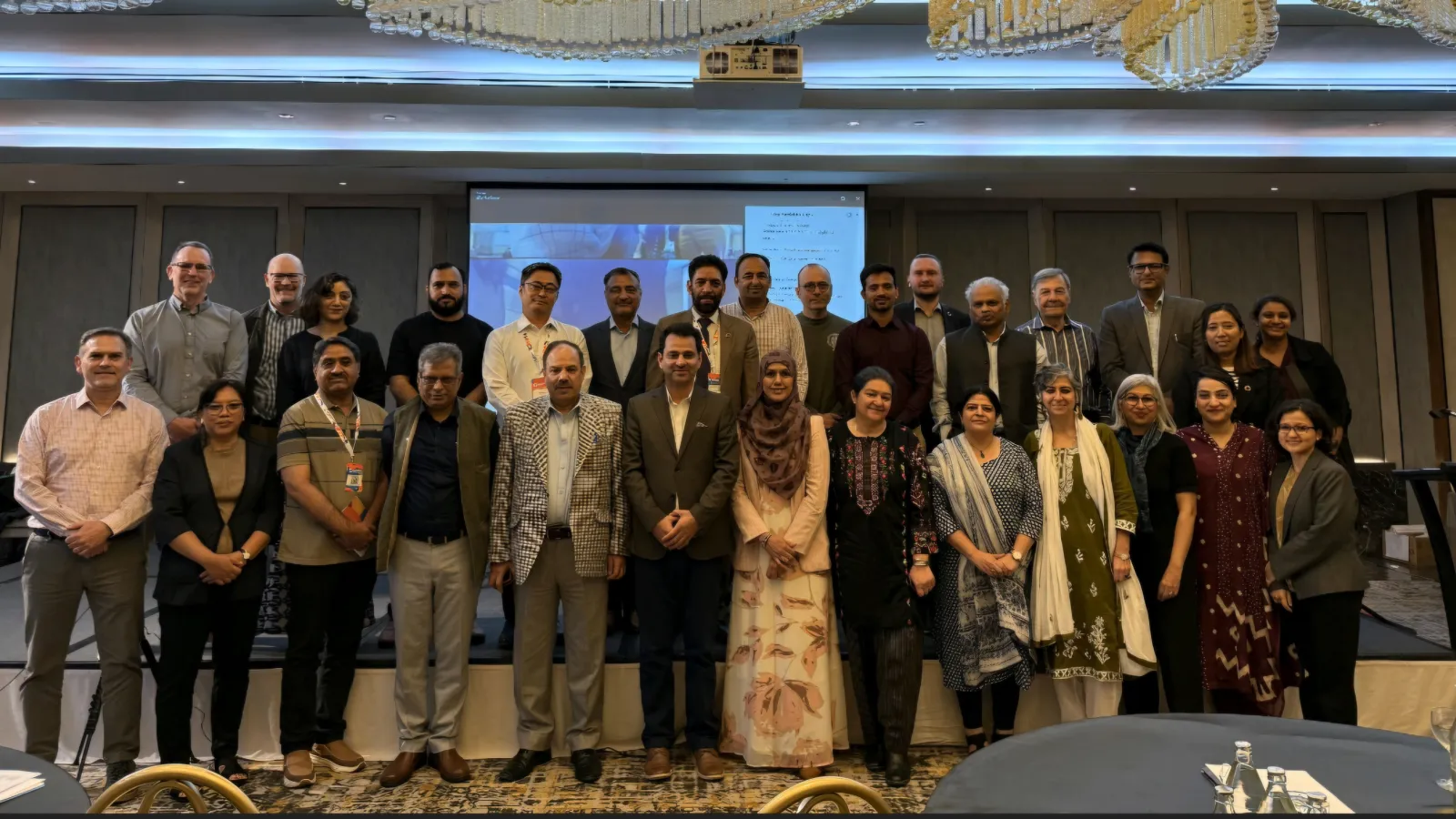 Group photo of about two dozen workshop participants standing on a stage in a conference room, with a projection screen behind them.