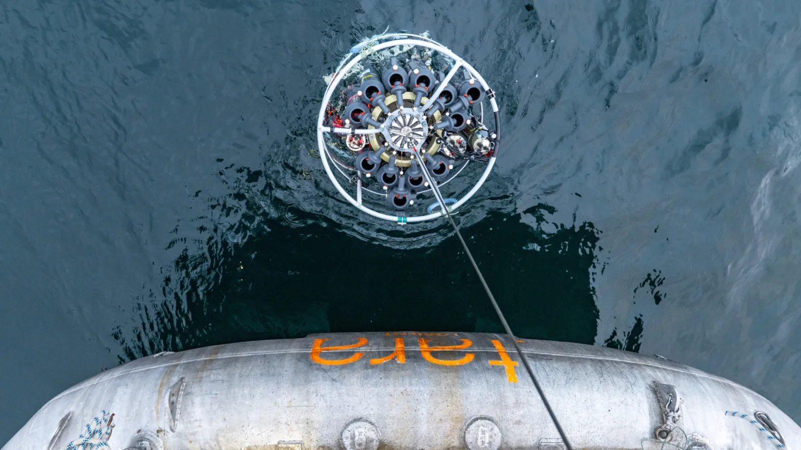 Overhead view of a circular ocean-sampling instrument array being lowered into the sea from a ship.