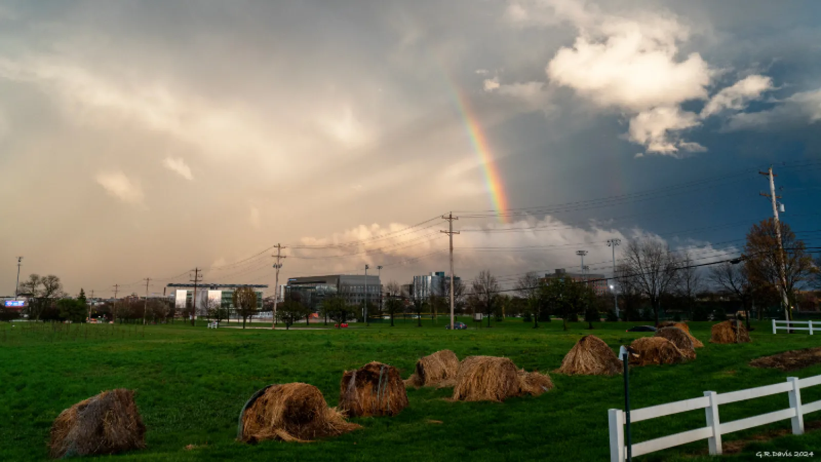 A green field with haystacks sits under stormy skies and a bright rainbow, with houses and power lines in the distance, a white fence on the right, and the signature “G.R. Davis 2021.”