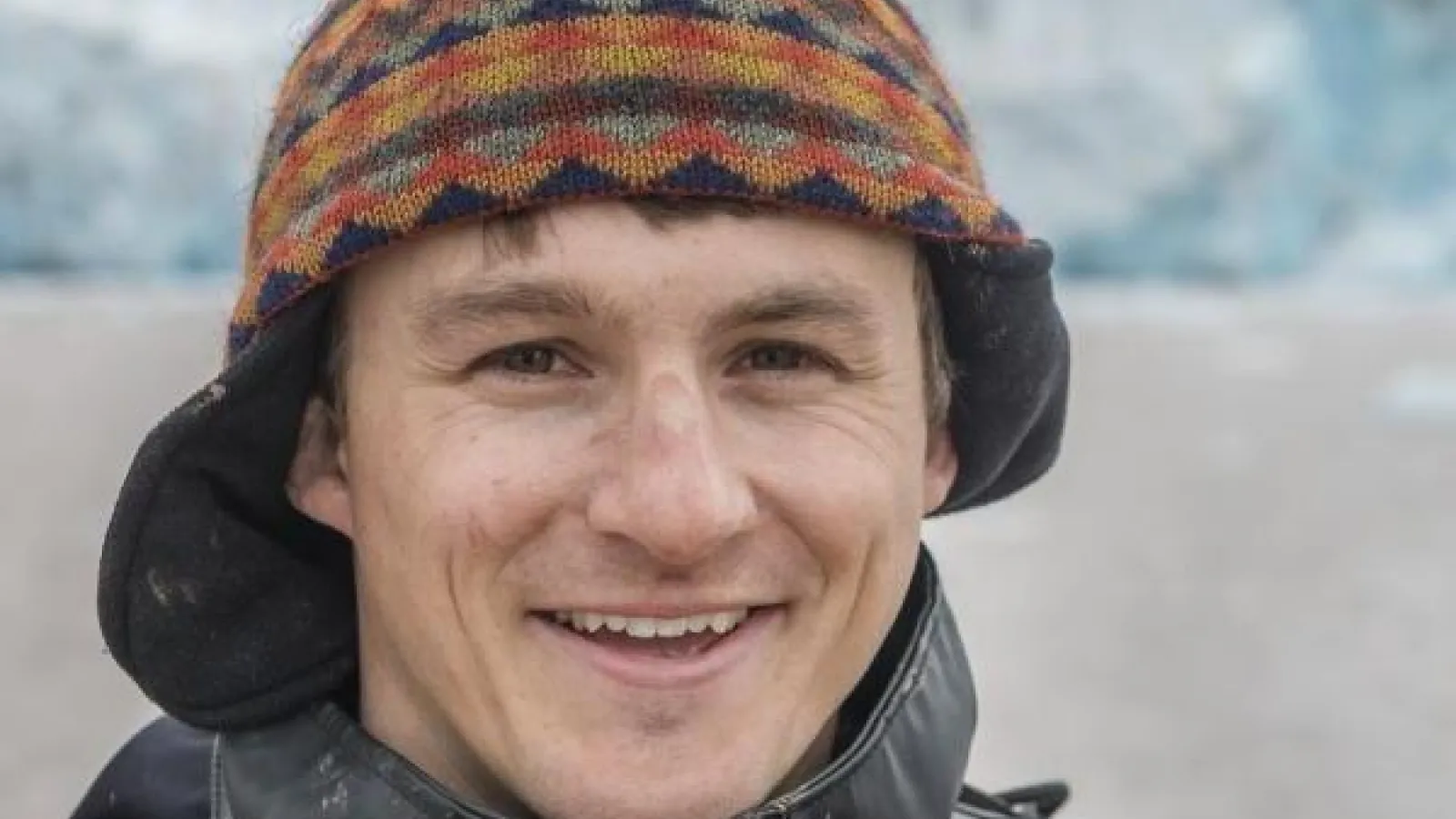 Close-up of Alex Michaud smilingwearing a colorful knit hat and a black jacket, with a blurred icy outdoor background.