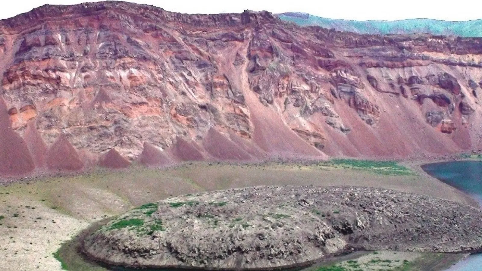Colorful layered cliffs with red, purple, and tan sediment slopes descending to a rocky shoreline beside a blue lake, with sparse green vegetation at the base.