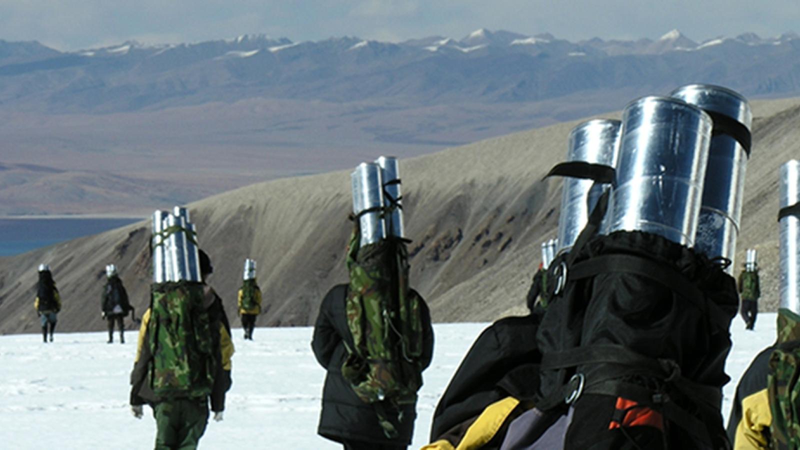 A landscape of snow covered ground and brown mountains in a distance and multiple people walking away with metal cylinders in their backpacks.