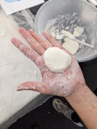 A white, dough ball in the palm of a hand with a bowl on a table in the background.