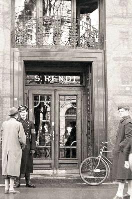 A black and white photo of the front doors of Tuchhaus S Rendi. The doors are gilded, the street is clean, there is one civilian crossing into frame on the right and another speaking to a Nazi officer on the left. They are all dressed in long coats and hats.