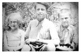 Three children, a girl and two boys, sitting in front of some plants. The girl and boy on the right are smiling, the boy on the left is grimmacing in the sun.