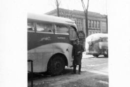A vintage greyhound bus with a man in a coat standing outside its door. The bus is in a parking lot with a building in the background.