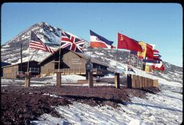 A building on the top of a mountain labeled "National Science Foundation." In front are a dozen flags representing countries from all around the world including the United States, the United Kingdom, Communist China, Norway, Japan, Brazil, Germany, Australia, and others that are not fully visable. The mountain is covered in snow.