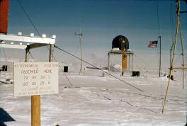 Sign with astronomic position at Byrd Station sitting on the left of the image. The writing is red on a white plaque on a wooden post. In the background are various structures and ropes. With a water tower-like structure with an American flag on a flagpole.