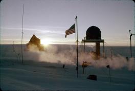 The sun at the horizon illuminates the image. In the forefront is an American flag on a flagpole. In the middle ground are various structures including a raised dome and a canvas covered space.