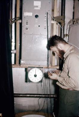 Brecher is standing to the right with a tool and working on a wire of the spectrograph. The spectrograph is a large metal box with about 10 different size knobs in the middle. At the bottom is an analogue timer.