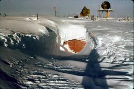 In the forefront of the image is an orange metal garage door visible in the snow. It lays below the surface. In the background of the image are poles and structures to help maintain the camp below.