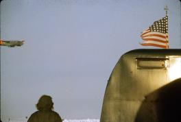 In the upper left corner is a dark colored airplane with red tips. On the right side of the image is a tattered American flag on a pole and a piece of metal, perhaps a door, underneath that. There is a figure to the left of this looking up at the plane with their hood up. The sky is blue in the background.