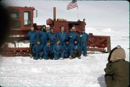 Traverse party is posing in front of the side of the red tractor train front. There are 6 men standing and another 5 kneeling in front of them. All are wearing blue snow suits. The tractor train has the American flag attached to the front. There is another person in brown in the foreground taking a picture of the posed group of men. There is snow on the ground.