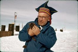 Havola is in a blue jacket and holding a black kitten in his hands looking at the camera. Behind Havola are fuel barrels scattered around the snow.