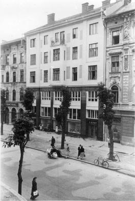 A view of Tuchhaus S. Rendi from across the street. The whole five stories are in view and people can be seen on the street below walking and looking into the windows of the shop