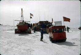 Three red vehicles are preparing to leave the station. The first is a small red car in the front with a red flag on the side. The second is to the left is a medium size snow truck with a large crane at the top. The largest is the snow tractor train behind the small truck. The large tractor train has multiple carts attached. There are 3 people in between these vehicles attending to various tasks.