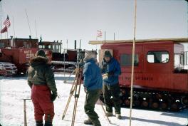 Three individuals are in snowsuits outside on the snow. Two have their backs to the camera and the other one is writing something down on a pad of paper. The man in the middle is looking through a tripod. On the right side is a red snow truck is wood on top. The left has a row of red snow trucks further back with American flags on them.