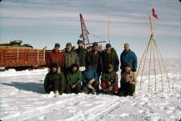 Traverse party posing in front of a bamboo stick marker. The sticks are in line and tied together at the top. These 11 men are all wearing jackets, gloves, hats, and other winter wear. Half are standing and the other half kneeling in front of them. In the background is part of a tractor train. The visible part are pens of red wood with material inside.