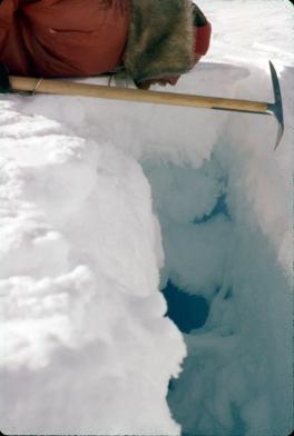 Forrest Dowling peering into crevasse. Dowling is laying on his stomach in the snow with a pic ax in his hand. He is wearing a red coat with a fur hat. Dowling is looking down into a large crevasse where you can see both water and ice.