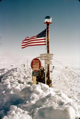 Signs at South Pole Station entrance standing out in the snow. The flagpole has the American flag hanging out it. It is decorated in multiple colors with a clear globe at the top. At the bottom of the pole is a small pine tree next to a few signs that point in different directions. These signs have names of cities and their distance from the location.