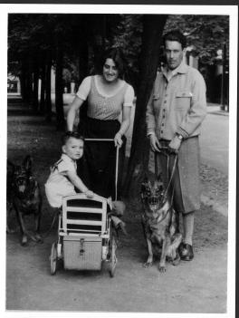A balck and white photo of a toddler in a stroller being pushed by his mother with his father and two dogs standing next to them