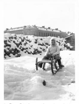 A young child in a sled on a hill with a large building behind him in the distance.