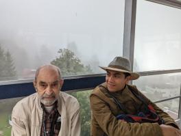 Henry, an old man in a wind breaker and a flanel, sitting next to Gabriel, a young man in a thick coat and broad brimed hat. They are sitting in a class box. There are trees and grass far below them.