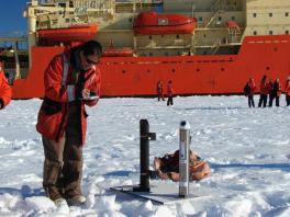 A white woman in a red coat standing in a field of ice with a large boat and people milling about behind her. She writes in a notebook and stands next to a pair of scientific instruments.