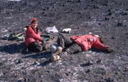 Two white women in thick red coats hanging out in a dry rock valley. The one on the left is sprawled on the ground, the one on the right is sitting up and smiling.