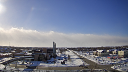 snow covered rural city scape with some buildings and streets, some snow covered areas under sunny blue skies and white clouds  