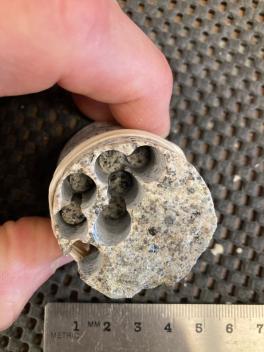 Someone holding a small 6mm diameter core toward the cameral with smaller drill holes inside of it. Backdrop is a porous mat and a silver ruler.