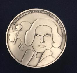 Lois Jones Medal Front. It is a gold medalion engraved with the head and shoulder of a woman with a bob haircut and a coat. Beside her is an aerial view of the Antarctican continent. On the top lip of the medallion, the full name of the award has been engraved.