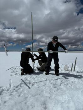 Three people drill into snow on a glacier under cloudy skies
