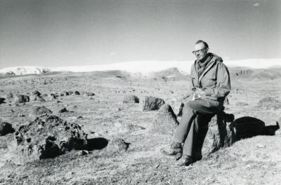 Black and white photo of man with glasses and long jacket sitting on a rock  in a rocky field with snow and ice in a distance
