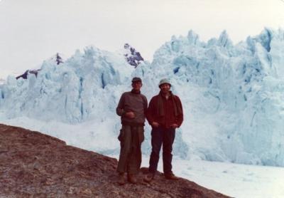 Two men dressed for cold weather standing on a rocky hill in front of a huge pile of ice and snow